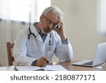 Stressed middle aged old family doctor general practitioner in white uniform and eyeglasses working alone in clinic office, thinking of problems solution or involved in medical paperwork indoors.