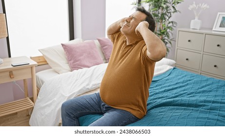 A stressed middle-aged man sitting on a bed in a bedroom, expressing a headache or frustration with a hand on his head. - Powered by Shutterstock