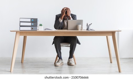 Stressed millennial black businessman using laptop, sitting at desk with head in his hands, making mistake in business project, failing to meet deadline against white studio wall, full length