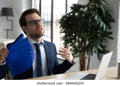 Stressed Millennial Businessman In Formal Suit And Eyewear Feeling Overheated In Office Working Indoors Without Air Conditioning System, Unhappy Male Manager Using Paper Fan Blowing Fresh Air.