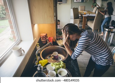 Stressed Mum At Home. She Has Her Head In Her Hands At A Messy Kitchen Sink And Her Children Are Running Round In The Background.