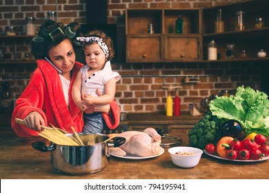 Stressed Mum At Home. Young Mother With Little Child In The Home Kitchen. Woman Doing Many Tasks While Looks After Her Baby