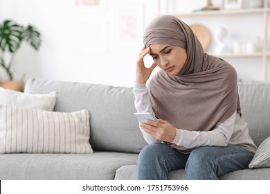 Stressed Muslim Woman In Hijab Waiting For Important Call At Home, Sitting On Couch With Cellphone In Hands, Free Space - Powered by Shutterstock