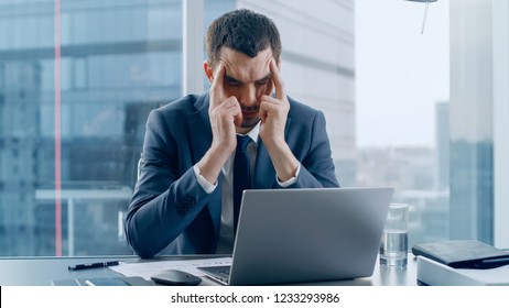 Stressed Out Businessman Sitting at His Desk, Massages His Head in Frustration. Trying to Concentrate. Overworked Businesspeople In the Window Big City Business District View.
