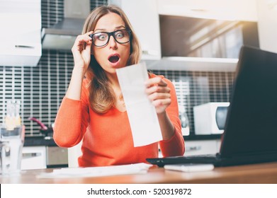 Stressed Over Bills. Portrait Of Surprised Young  Woman Using A Laptop Computer Sitting At Her Kitchen Holding Utility Bill And Bank Statements. Home Interior.