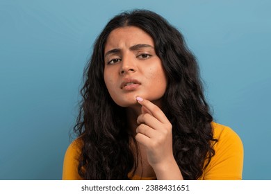 Stressed sad brunette young indian woman touching her face over blue studio background, having skin problems, closeup. Acne, pimples, wrinkles, dull skin
