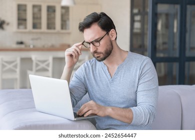 Stressed sad tired serious young caucasian man businessman student professor freelancer working using laptop sitting on the sofa at home indoors. Home office, business problems, remote work - Powered by Shutterstock