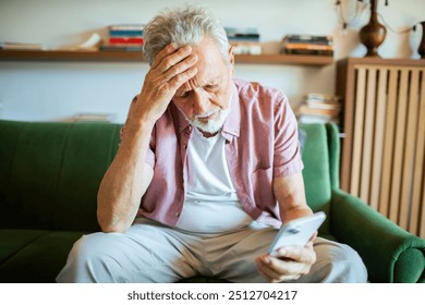 Stressed senior man holding head and smartphone on home sofa - Powered by Shutterstock