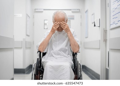 Stressed Senior Patient Sitting On Wheelchair In Hospital Corridor. Lonely Old Man Cover His Eyes With Hands In Hospital Hallway. Elderly Disabled Patient Feeling Sad And Abandoned.