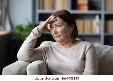 Stressed senior retired woman touching forehead, looking away, feeling doubtful about decision. Unhappy thoughtful middle aged lady sitting on couch, worrying about personal problems alone at home.