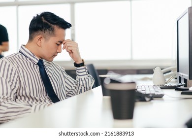 Stressed Serious Asian Male Employee Feel Pain Eyestrain Working On Computer. Tired Man Suffer From Headache Bad Vision Sight Problem Or Company Financial Crisis At Desk In The Office.