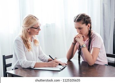 Stressed Teenage Girl Sitting At Table On Therapy Session By Female Counselor Writing In Clipboard In Office 