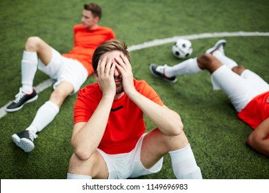 Stressed or tired footballer sitting on green field and covering his face with hands on background of two mates - Powered by Shutterstock