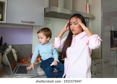 Stressed And Tired Mother Having Headaches While Working From Home With Her Kid On Messy Kitchen. Young Asian Woman Housewife Overwork With Annoying Child.