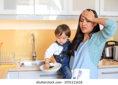 Stressed And Tired Mother Suffering From Headaches While Doing Housework With Her Kid. Asian Woman Cleaning Dishes With One Hand Carrying Her Baby At Kitchen Home. Housewife Overwork Concept.