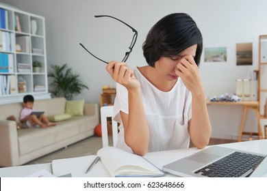 Stressed Tired Woman At Her Workplace, Her Child Sitting On Sofa Behind Her