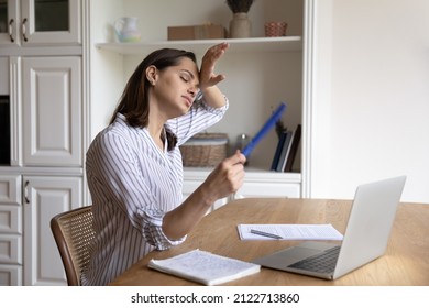 Stressed Unhappy Young Businesswoman Waving Paper Fan, Blowing Cooled Air, Suffering From Hot Temperature In Modern Home Office Without Air Conditioner, Feeling Overheated And Exhausted Alone Indoors.