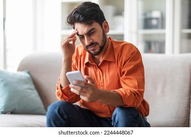 Stressed upset arab man in casual sitting on couch at home, holding cell phone, looking at gadget screen and touching his head, reading bad news, reading message, copy space