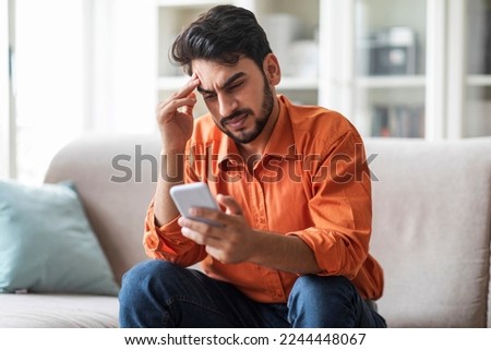 Stressed upset arab man in casual sitting on couch at home, holding cell phone, looking at gadget screen and touching his head, reading bad news, reading message, copy space