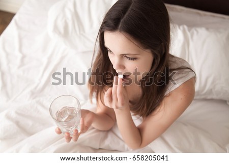 Stressed woman drinking white round pill while sitting in bed with glass of water in hand. Tired young lady takes medicines after wake up in the morning, tries to overcome insomnia with sleeping pill Foto d'archivio © 