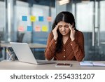 A stressed woman in an office setting, feeling a headache while working on a laptop with a smartphone nearby. She