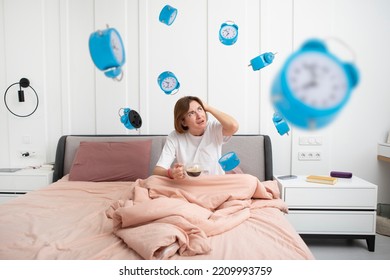 Stressed Woman Sitting On The Bed With A Cup Of Coffee And Looking At Lots Of Alarm Clocks Flying Around Her. Wake Up Early And Don't Sleep Concept 