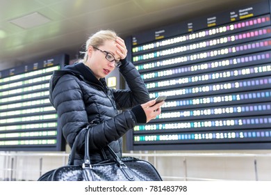 Stressed Worried Woman In International Airport Looking At Smart Phone App Information And Flight Information Board, Checking Her Flight Detailes.