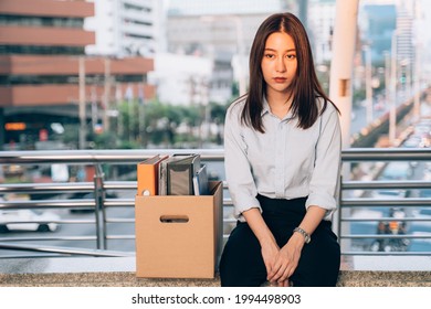 Stressed And Worried Young Asian Woman With Box Of Items Sitting Alone After Being Laid Off From Job Due To Recession And Economic Stress In Industry