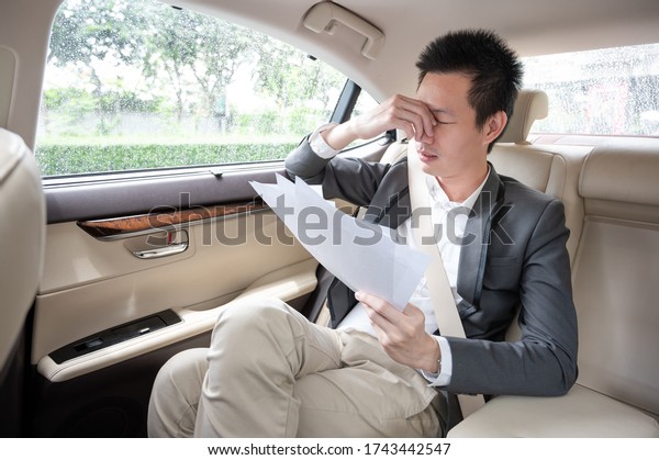 Stressed young asian\
businessman looking at Financial documents with expression Tired\
and worried while sitting on the back seat in the car.  many bad\
financial report.