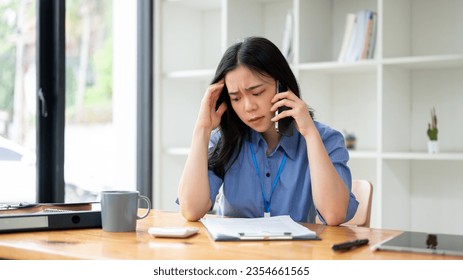 A stressed young Asian businesswoman is feeling overwhelmed, trying to fix the problems over the phone, and having a serious conversation over the phone while sitting at her desk in the office. - Powered by Shutterstock