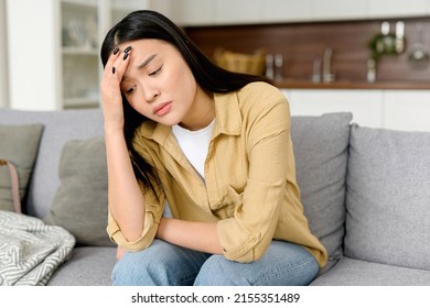 Stressed young asian woman touching head and feels dizzy, suffering from headache and migraine pain, sits on the sofa at home, tired and exhausted lady feels bad - Shutterstock ID 2155351489