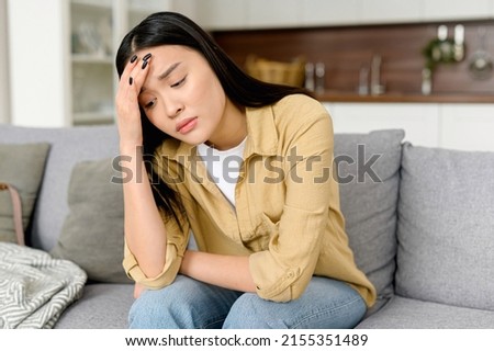 Stressed young asian woman touching head and feels dizzy, suffering from headache and migraine pain, sits on the sofa at home, tired and exhausted lady feels bad