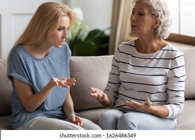 Stressed young blonde grown up daughter arguing with nervous old mature mother, sitting together at home. Irritated elderly woman lecturing adult child, different generations misunderstanding gap.