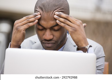 stressed young businessman sitting outside corporate office working on laptop computer holding head with hands looking down. Negative human emotion facial expression feelings.