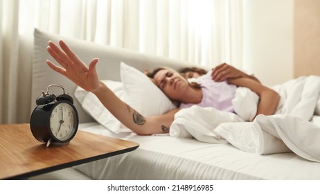 Stressed young european woman pulling hand to stopping alarm clock while sleeping with boyfriend under blanket on bed at morning. Domestic lifestyle. Idea of rest at home. Bedroom in spacious flat