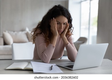 Stressed young hispanic woman looking at computer screen, stack with difficult task, feeling confused getting email with bad news, suffering from headache due to long work on online project at home.