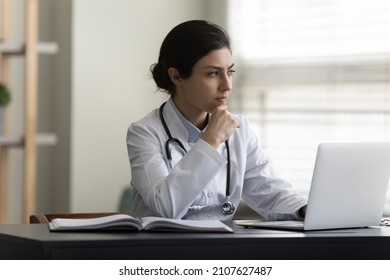 Stressed Young Indian Female Doctor Therapist In White Medical Uniform Looking In Distance, Thinking Of Difficult Problem Solution, Considering Illness Treatment Working On Computer In Modern Clinic.