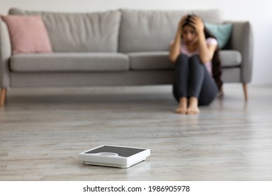 Stressed young Indian woman sitting on floor, grabbing her head in terror, looking at empty scales, feeling overweight, selective focus. Copy space. Weight loss, eating disorder, anorexia concept