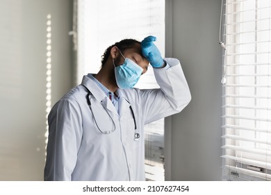 Stressed Young Male African American Doctor Physician In Protective Gloves And Disposable Facemask Wiping Sweat From Forehead, Feeling Exhausted After Hard Working Day In Infectious Department.