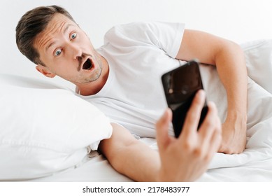 Stressed young man with emotional expression and open mouth holding phone and looking at time of alarm clock on smartphone while lying on bed in bedroom. Wake up shocked guy late for work in morning. - Powered by Shutterstock