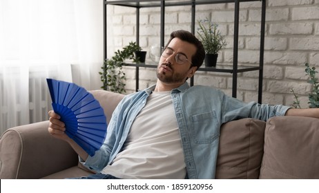 Stressed Young Man In Glasses Sitting On Sofa, Suffering From Overheating Or Hot Summer Weather Without Air Conditioner, Using Paper Fan Cooling Himself, Breathing Fresh Air, Relaxing Alone At Home.