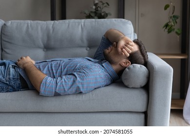 Stressed young man lying on sofa on pillow, covering face with hand, suffering from negative thoughts or insomnia, having lack of energy, sleeping daydreaming napping at home, recreation concept.