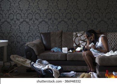 Stressed Young Mother Sitting On Her Sofa Whilst Feeding Her Baby Son. She Has Her Head In Her Hand And Is Surrounded By Mess.