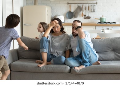 Stressed Young Parents Sitting On Couch, Tired Of Crazy Loud Hyperactive Little Children Siblings At Home. Unhappy Married Couple Irritated By Naughty Small Kid Son Daughter, Suffering From Headache.