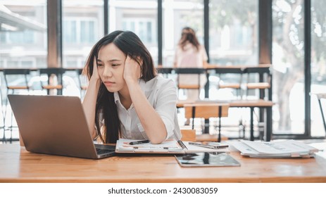 A stressed young woman holds her head in frustration while working at her laptop in a cluttered office environment. - Powered by Shutterstock