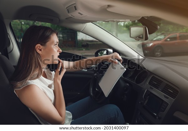 Stressed young woman with notebook talking on\
phone in driver\'s seat of modern\
car