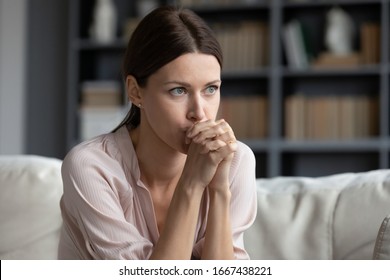 Stressed young woman sitting on couch, worrying about problems alone at home, afraid of difficulties, looking away, head shot. Angry nervous frustrated brunette lady sitting on sofa, feeling jealous.