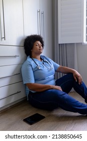 Stressful African American Mid Adult Female Nurse With Eyes Closed Sitting On Floor In Hospital. Unaltered, Healthcare Worker, Stress, Expertise And Occupation Concept.