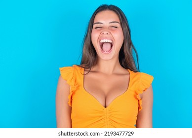 Stressful Beautiful Brunette Woman Wearing Orange Tank Top Over Blue Background Screams In Panic, Closes Eyes In Terror, Keeps Hands On Head, Finds Out Terrified News, Can't Believe It.