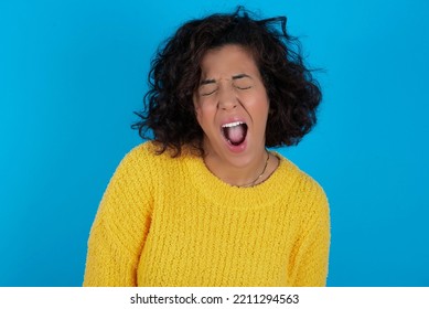 Stressful Brunette Arab Woman Wearing Yellow Sweater Over Blue Background Screams In Panic, Closes Eyes In Terror, Keeps Hands On Head, Finds Out Terrified News, Can't Believe It.